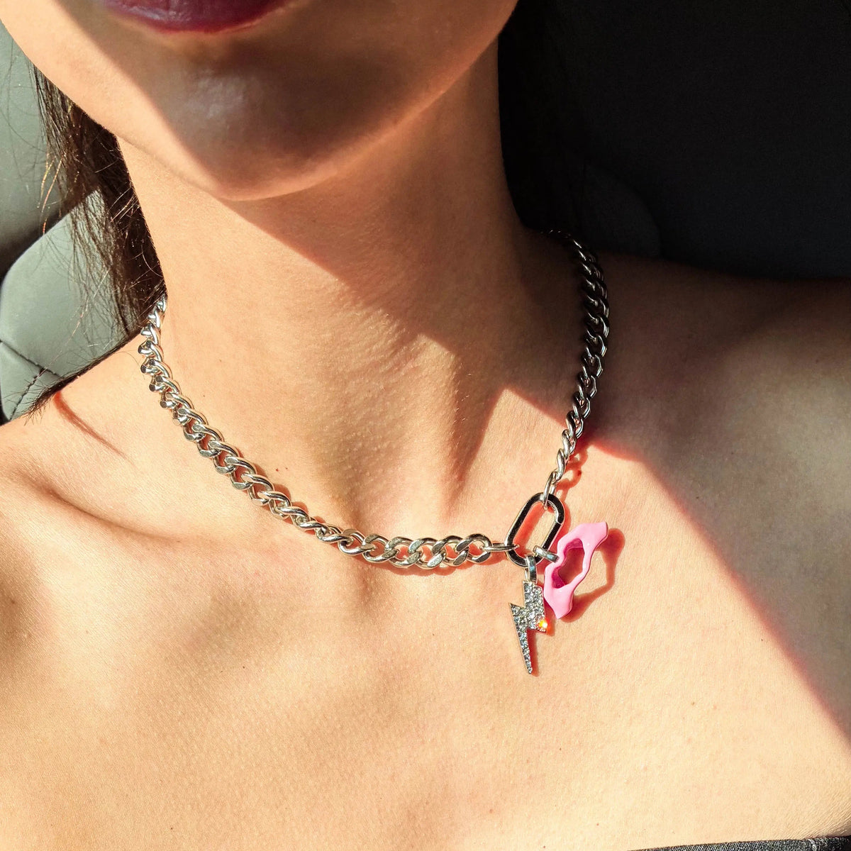 A close-up of a woman wearing a silver chain necklace with a pink heart-shaped charm and a small silver lightning bolt detail